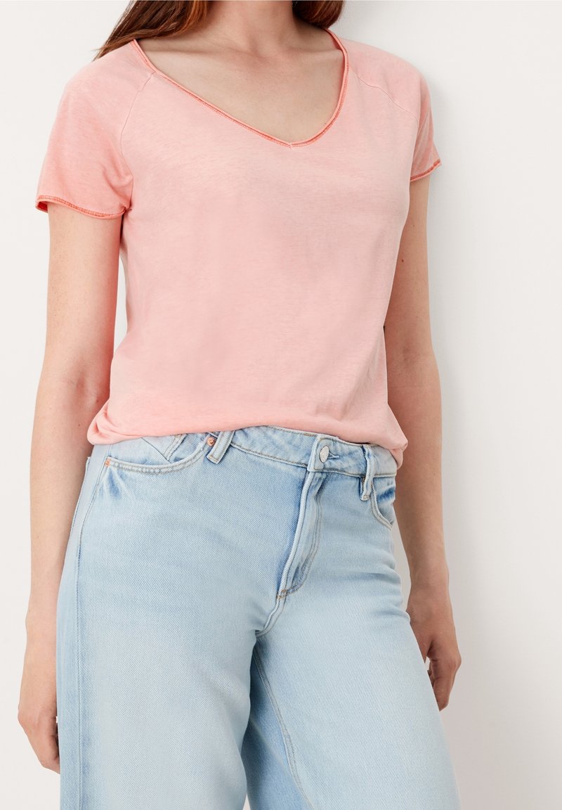 Femme portant un T-shirt rose clair à manches courtes et col en V, rentré dans un jean en denim bleu clair taille haute, debout contre un mur blanc.