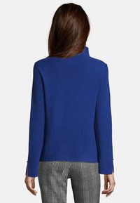 Kvinna med långt brunt hår som bär en blå texturerad långärmad jacka och svartspräckliga byxor, visad bakifrån.