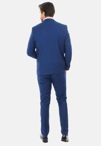 Abito su misura blu con rever a punta, spacco singolo sul retro e pantaloni slim-fit, abbinato a scarpe nere lucide.