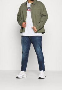 Olivově zelená košile s dlouhým rukávem a dvěma náprsními kapsami, šedé tričko, tmavě modré džíny a bílé tenisky s černými detaily.