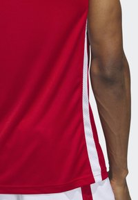 Rotes Sporttrikot mit weißen Mesh-Seitenpaneelen, hat eine glatte Textur und einen ärmellosen Schnitt. Sichtbare Nähte am Saum.