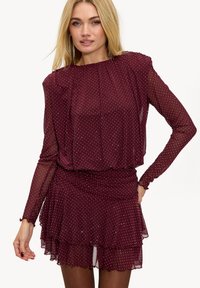 Robe bordeaux à pois avec un ourlet à volants superposés, des manches longues en voile transparent et un col froncé, en tissu léger et fluide.