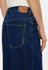 Σκούρο μπλε τζιν παντελόνι με καφέ δερμάτινη ετικέτα από κομμένο κείμενο. Ορατές ραφές και δύο πίσω τσέπες με διακοσμητικές ραφές.