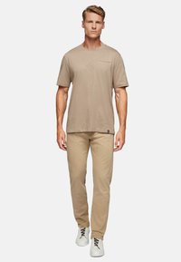 Men's beige cotton t-shirt with a crew neck and a small chest pocket, paired with light beige trousers and white shoes with dark laces.