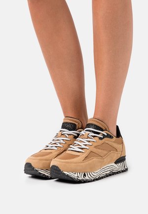 Nohy v béžových semišových teniskách s bílými tkaničkami a podrážkami s černobílým vzorem na bílém pozadí.