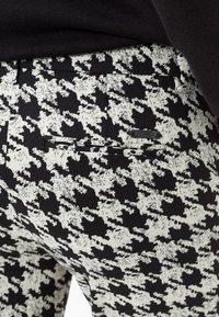 Pantalon taille haute à motif pied-de-poule noir et blanc avec une texture lisse, doté d'une poche arrière avec une fermeture à bouton noir.