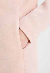Prenda de color rosa claro con un acabado texturizado, que cuenta con un bolsillo lateral y un corte en la manga, destacando un diseño suave y casual.