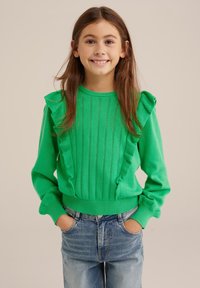Pulover verde strălucitor cu umeri volantați, model texturat de tricot și mansete cu nervuri. Asortat cu blugi din denim de culoare albastră deschisă.