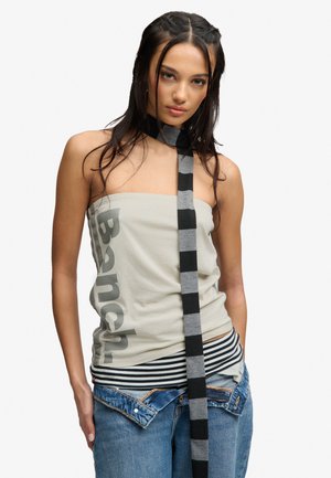 Ung kvinde med fletninger bærer en skulderfri top med "Bench" logo, stribet taljedetalje, løstsiddende lavtaljede jeans og et langt sort- og gråstribet halstørklæde.