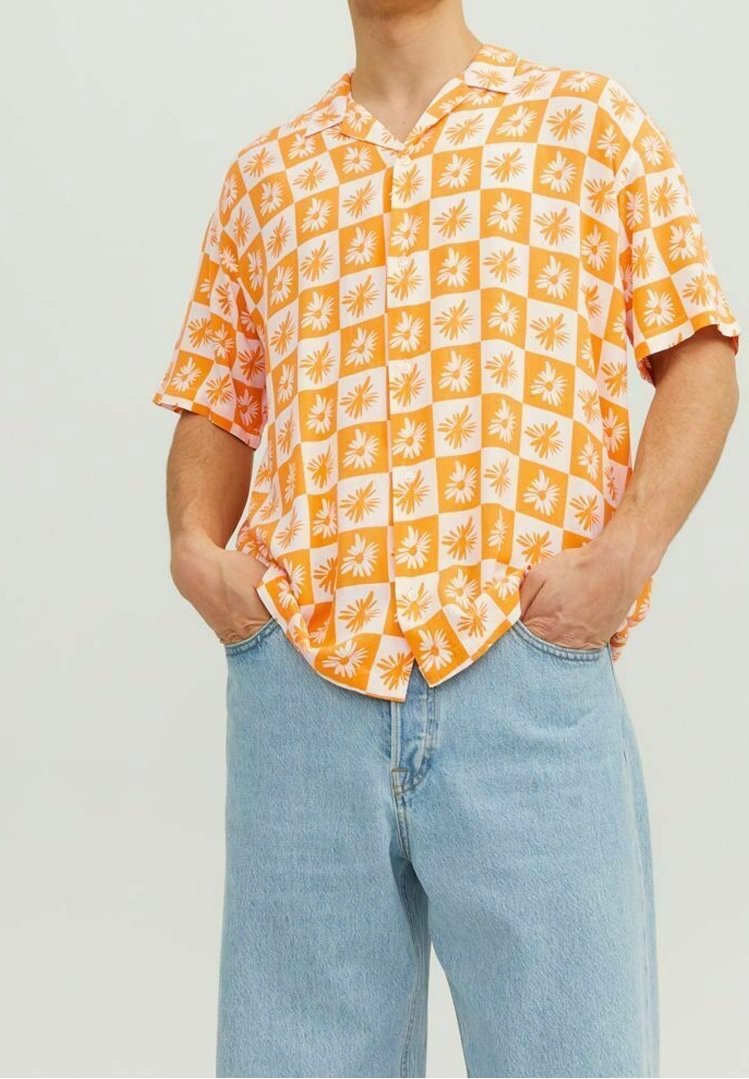 Homme portant une chemise à manches courtes à carreaux orange et blancs avec un motif floral, associée à un jean en denim bleu clair. Mains dans les poches.