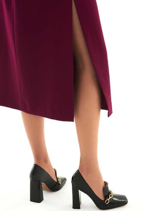Robe bordeaux avec une fente latérale ; chaussures à talons hauts noires vernies avec un détail de chaîne dorée. Texture lisse et forme angulaire visibles.