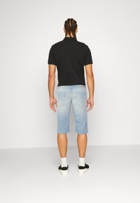 Camisa polo negra, pantalones cortos de denim azul claro y zapatillas negras con suelas blancas. Modelo de espaldas, mostrando la vista trasera del atuendo.