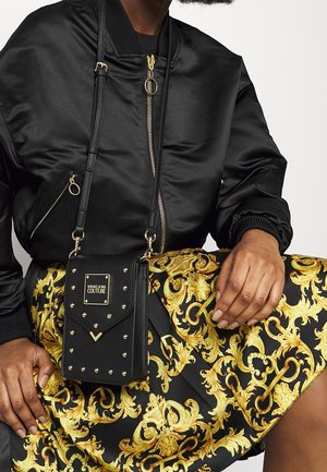 Borsa a tracolla nera con borchie dorate e una targa, abbinata a una gonna con un motivo barocco in nero e oro. Giacca bomber che valorizza il look.