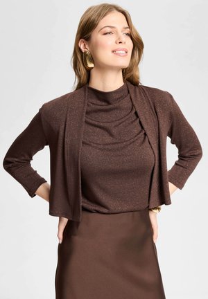 Kvinde smiler med hænderne på hoften, iført skinnende brun top, matchende brun cardigan, nederdel, store guld hoop øreringe og et guldt ur.