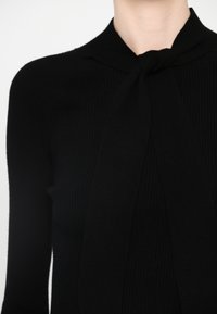 Black ribbed knit top with a tie detail at the collar, featuring long sleeves and a textured surface for a fitted look.