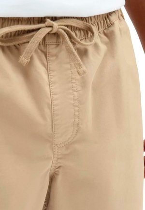 Pantalones beige con cinturilla elástica y cordón ajustable. Cuenta con bolsillos laterales y costuras cosidas, mostrando un tejido ligero y suave.