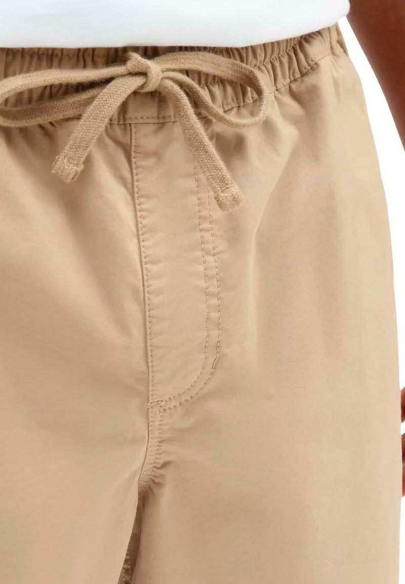 Pantalons beiges avec une ceinture élastique et un cordon de serrage. Dotés de poches latérales et de coutures surpiquées, mettant en valeur un tissu lisse et léger.