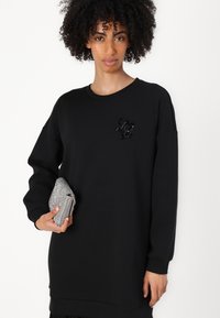 Sweatshirt noir à coupe ample, avec des manches longues et un col rond. Présente un logo texturé et perlé sur la poitrine. Tient une pochette argentée.