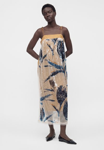 Femme portant une robe beige sans manches avec des sequins floraux bleus et noirs, debout les mains derrière le dos et chaussures plates noires pointues.