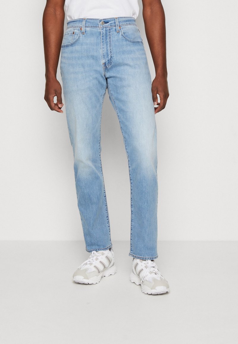 Homme portant un jean droit bleu clair et des baskets blanches, debout devant un fond uni.