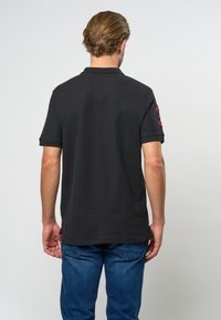 Černé polo tričko s krátkými rukávy, strukturovanou látkou, límečkem a malými červenými akcenty na rukávech. Nosit s modrými džínami.