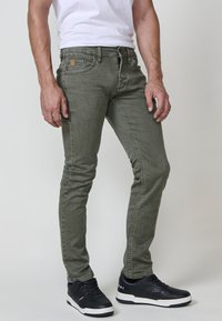 Olivno zeleni skinny jeans s klasičnim petim žepom, šivnimi detajli in gumbom, v kombinaciji s črnimi supergami.