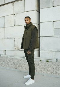 Gilet matelassé vert olive porté sur un pull sombre, associé à un pantalon cargo olive et des baskets blanches. Tissu texturé et coupe ajustée.