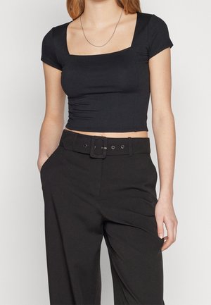 Femme portant un crop top noir ajusté avec un col carré, associé à un pantalon noir taille haute et une large ceinture noire avec une boucle carrée.