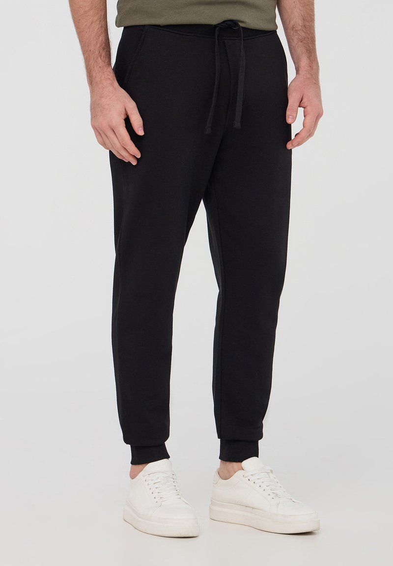 Czarne spodnie dresowe wykonane z miękkiego materiału, z elastycznym pasem wciąganym z sznurkiem, zwężanymi nogawkami i prążkowanymi mankietami. Zestawione z białymi sneakerami.
