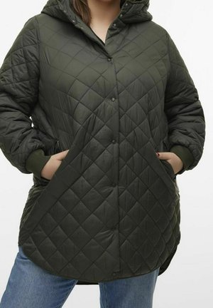 Femme portant une veste matelassée à capuche vert foncé avec boutons-pression, les mains dans les poches, associée à un jean bleu.