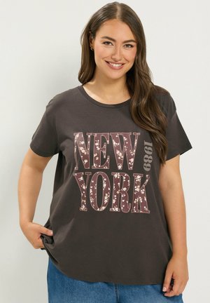 Uśmiechnięta kobieta z długimi brązowymi włosami, ubrana w czarną koszulkę z "NEW YORK 1989" i kwiatowymi wzorami, w połączeniu z niebieskimi dżinsami.