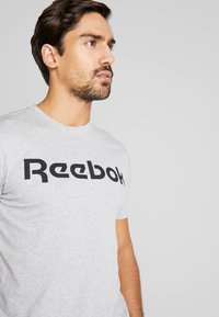 Grå t-skjorte med korte ermer, med en fremtredende svart "Reebok"-logo på brystet, laget av mykt bomullsstoff.