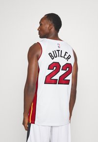 Balts basketbola krekls ar sarkaniem un zeltainiem akcentiem, kur aizmugurē ir virsraksts "BUTLER" un numurs "22" treknā melnā krāsā.