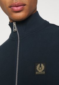 Marinblå zip-jacka med hög krage, mjukt tyg, med en guld- och svart broderad logotyp på bröstet och metallicdetalj på dragkedjan.