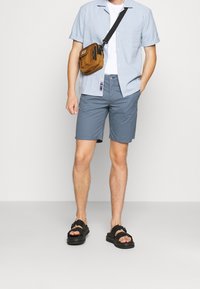 Camisa de rayas azul y blanca de manga corta, pantalones cortos grises, sandalias negras con correas, bolso cruzado marrón y una camiseta blanca debajo.