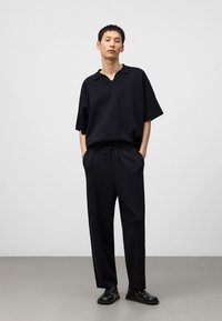 Zwarte oversized polo shirt met een uitgesneden kraag, gecombineerd met losse zwarte broeken met een trekkoord in de taille. Draagt zwart schoeisel.