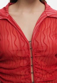 Gros plan d'une personne portant un haut rouge transparent à motifs ondulés, partiellement fermé avec des fermetures à crochets noires.