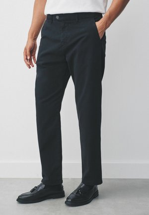 Black chinos featuring a button closure, side pockets, and a tapered fit. Smooth texture with a slight sheen, styled with black loafers.