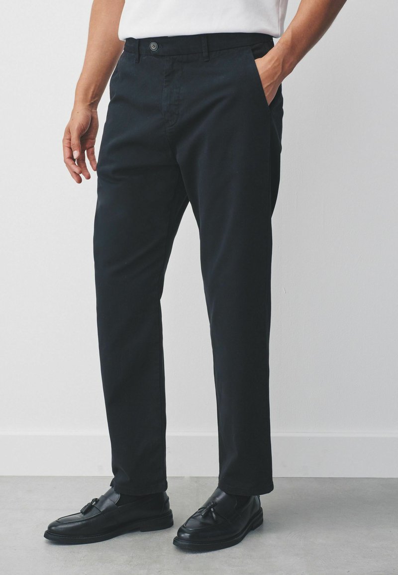 Black chinos featuring a button closure, side pockets, and a tapered fit. Smooth texture with a slight sheen, styled with black loafers.