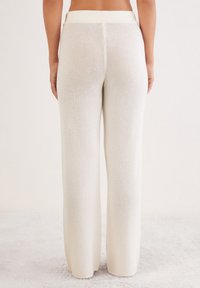 Femme debout pieds nus sur un tapis clair, portant un pantalon en tricot couleur crème, taille haute et jambes larges, vu de dos.