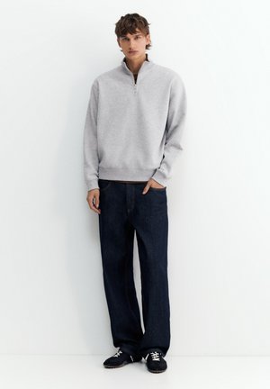 Jeune homme debout portant un sweat-shirt gris clair à fermeture éclair quart, un jean bleu foncé et des baskets noires sur un fond blanc.