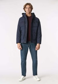 Giacca imbottita blu navy con cappuccio, dotata di quattro tasche frontali e chiusura con zip, indossata sopra una camicia bordeaux e jeans blu, abbinata a sneakers bianche.