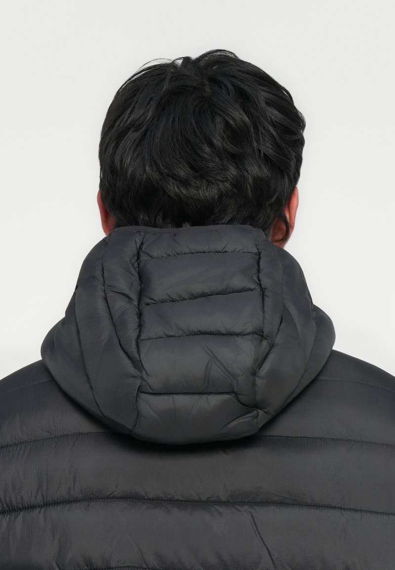 Man met kort zwart haar, gekleed in een zwart gevoerd jack met een dikke capuchon, gezien van achteren tegen een effen achtergrond.