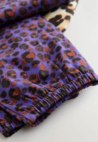 Fialová látka s leopardím vzorem v hnědé a černé barvě, s elastickým lemem. Hladká textura a živé barvy jsou výrazné.