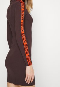 Femme portant une robe ajustée côtelée de couleur marron foncé avec une bande rouge sur la manche comportant le texte répétitif "KORS".