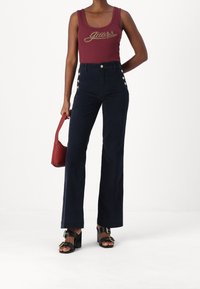 Mujer vistiendo una camiseta sin mangas burdeos de "Guess", pantalones de tiro alto en navy con botones laterales, sandalias de tacón negras y sosteniendo un pequeño bolso rojo.