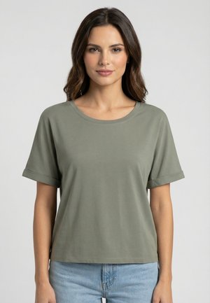 Mujer con cabello castaño hasta los hombros, vistiendo una camiseta de manga corta lisa color verde oliva y jeans azul claro, de pie frente a un fondo sencillo.