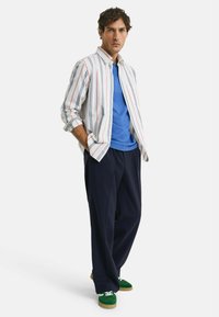 Pruhovaná košile na knoflíky v bílé, červené a modré barvě přes modré tričko. Volné námořnické kalhoty a zelené semišové tenisky doplňují outfit.