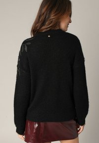 Pull noir avec un tricot texturé, présentant un motif de feuilles subtil sur l'épaule gauche et un petit accent doré au dos de l'encolure.