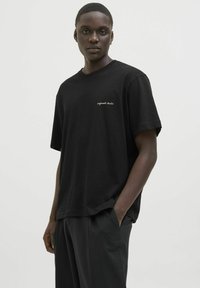 Sort bomulds t-shirt med en afslappet pasform, der har en lille hvid logo-tekst "original studio" på venstre bryst.